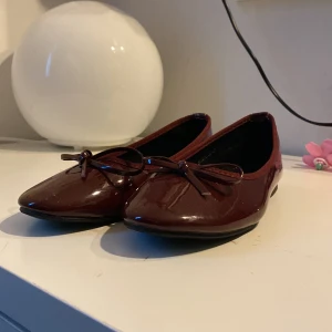 Vinröda ballerinaskor med rosett från Nelly - Säljer ett par glansiga vinröda ballerinaskor från Nelly med söt rosettdetalj framtill. Skorna har rund tå och platt sula, perfekta för en stilren och enkel look. 