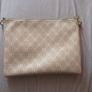 Beige kuvertväska från Don Donna - Säljer en beige kuvertväska från Don Donna med guldiga detaljer och dragkedja. Väskan har ett mönster med Don Donna-loggor över hela ytan och ett huvudfack med innerficka. Perfekt för att bära det viktigaste.