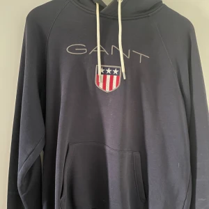 Svart hoodie från GANT med logga - Svart hoodie från GANT med broderad logga och sköld med stjärnor och ränder på bröstet. Tröjan har huva med vita snören och en stor magficka framtill. Perfekt för en avslappnad stil.