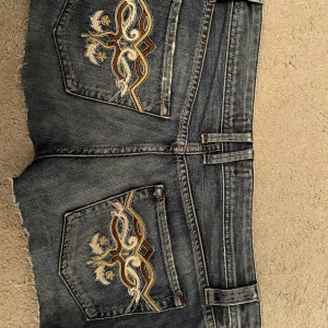 Jeansshorts med broderade bakfickor - Snygga blå jeansshorts med råa kanter och broderade detaljer på bakfickorna i vitt, gult och brunt. Klassisk femficksmodell med låg midja och slitningar framtill för en cool look.