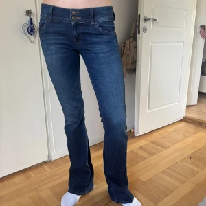 Mörkblå bootcut jeans med dubbla knappar - Säljer ett par mörkblå jeans med bootcut passform och dubbla knappar i midjan. Jeansen har snygga detaljer på bakfickorna med knappar och små flaggor. Klassisk femficksmodell och låg midja. Bra kvalite förutom att de är söndertrampade där nere vilket man kan se i sista bilden:)