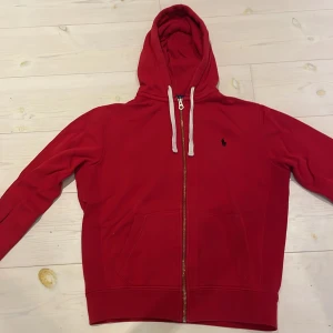 Röd hoodie från Polo Ralph Lauren - Röd hoodie med dragkedja från Polo Ralph Lauren. Klassisk design med huva och vita snören, två fickor framtill och broderad logga på bröstet. Perfekt för en avslappnad stil.