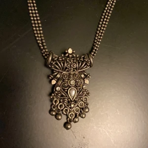 Halsband - Unikt halsband i antik silverfärg med dubbla kedjor och ett stort, detaljerat hänge med små kulor och ornament. Perfekt för dig som gillar bohemisk eller vintage stil. Säljs utan originalförpackning.