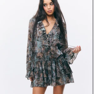 Mönstrad volangklänning från Monki - Supersöt kortklänning från Monki med volanger och v-ringning. Klänningen har ett unikt mönster i blått, grått och brunt, långa ärmar och knyt framtill. Perfekt för dig som gillar bohemisk stil och vill sticka ut.