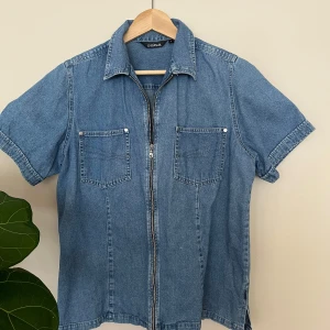 Jeansskjorta med dragkedja från Salsa - Säljer en klassisk blå jeansskjorta från Salsa med dragkedja framtill och två bröstfickor. Skjortan är gjord av stretchigt denimtyg, har korta ärmar och en rak passform. Perfekt för dig som gillar denim och vill ha en snygg-avslappnad stil.