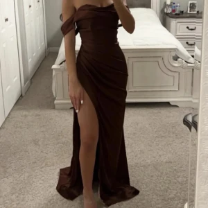 Balklänning - Säljer en elegant brun långklänning med offshoulder-design och hög slits framtill. Klänningen har en figurnära passform och draperade detaljer längs sidorna för en snygg siluett. Perfekt för bal, bröllop eller festliga tillfällen. Endast använd en gång