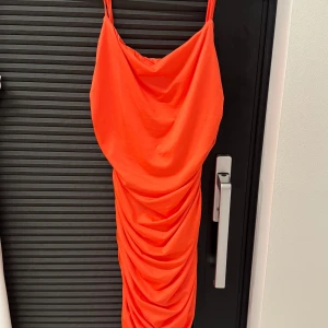 Orange draperad klänning med smala axelband - Säljer en snygg orange klänning med smala axelband och draperad effekt framtill. Klänningen har en figurnära passform och är tillverkad i ett mjukt, glansigt material. Perfekt för dig som vill sticka ut med en färgstark look.