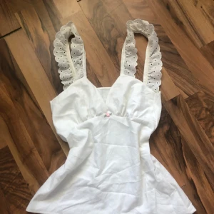 Vit linnetopp🌷 - Söt vit linnetopp med breda axelband i broderad spets och liten rosa ros framtill. Toppen har v-ringning och lätt utsvängd passform, perfekt för varma dagar.🌸 Lten fläck framtill☀️