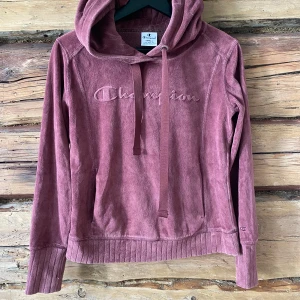 Rosa hoodie från Champion - Mysig hoodie i dammigt rosa från Champion med broderad logga på bröstet. Tröjan har huva med dragsko, magficka och ribbade muddar vid ärmslut och nederkant. Perfekt för en avslappnad stil.