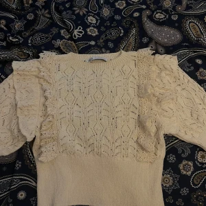 Beige spetsstickad tröja från Zara - Supersöt beige stickad tröja från Zara med spetsdetaljer och volanger längs axlarna. Tröjan har ett vackert hålmönster och ribbad nederkant. Perfekt för dig som gillar romantisk stil och vill ha något unikt i garderoben. Endast testad då den inte passade mig, dock ingen lapp kvar 🩷