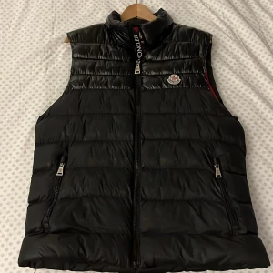 Svart dunväst från Moncler - Snygg svart dunväst från Moncler med hög krage och dragkedja framtill. Västen har två fickor med dragkedja och Moncler-logga på bröstet. Insidan är röd med unika Moncler-detaljer och etiketter. Pris kan diskuteras. Storlek M passar mer L. 