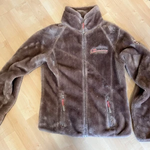 Lila fleecejacka från Nirvana 💜 - Mysig, varm fleecejacka från Nirvana med hög krage och dragkedja framtill. Jackan har två fickor med dragkedja och broderad logga på bröstet samt märke på ärmen. Perfekt för kyliga dagar.