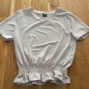 topp med volang - Säljer en söt vit ribbad topp från Gina Tricot i storlek XS. Toppen har korta ärmar, rund halsringning och en bred smock med volang nertill som ger en fin form. Perfekt till sommaren! Säljer pågrund av att jag har växt ut den.
