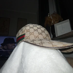 Äkta Beige keps från Gucci med GG-mönster - Säljer en äkta beige keps från Gucci med klassiskt GG-monogram och grönt/rött band på sidan. Justerbar rem baktill för perfekt passform. Kepsen är tillverkad i bomull och polyester, perfekt för dig som vill ha en lyxig accessoar. 