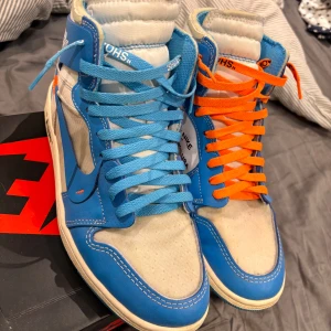 Nike Air Jordan 1 x Off-White UNC - Säljer ett par Nike Air Jordan 1 x Off-White UNC med blå och vita paneler, orange och blå skosnören samt Off-White-typiska detaljer och text på sidan. Skorna har hög modell och tydlig branding på hälen och tungan.