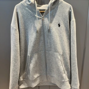 Grå hoodie med dragkedja från Ralph Lauren - Säljer en klassisk grå hoodie från Ralph Lauren med dragkedja och huva. Tröjan har två fickor framtill och den ikoniska svarta loggan broderad på bröstet. Perfekt för en avslappnad stil. Stl M