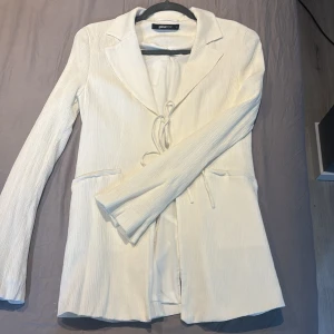 Set från Gina Tricot med knytning - Säljer en stilren vit kavaj med byxor från Gina Tricot. Kavajen har lång ärm, klassisk krage och är tillverkad i ett lätt, strukturerat tyg. Perfekt för dig som vill ha en fräsch och modern look. Har använt den en gång på studenten förra året. Kavajen är i storlek S och byxorna i storlek M. Nytt pris är 1200kr.