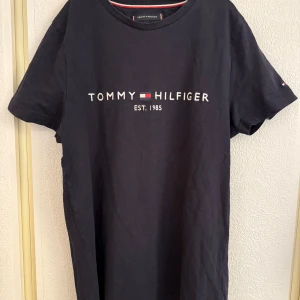 Mörkblå t-shirt från Tommy Hilfiger - Säljer en mörkblå t-shirt från Tommy Hilfiger med klassisk logga och texten 'EST. 1985' på bröstet. T-shirten är i ekologisk bomull och har rund hals och korta ärmar. Perfekt för en stilren och enkel look.
