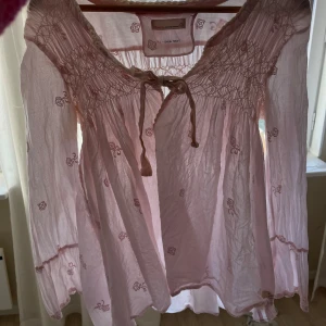 Ljusrosa blus med broderade rosor och knytband från odd Molly  - Supersöt ljusrosa blus från Odd Molly med broderade små rosor och smock upptill. Blusen har långa ärmar, volangdetaljer och knytband vid halsen. Perfekt för en romantisk look!