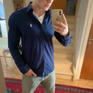 Mörkblå skjorta från Ralph Lauren - Säljer en stilren mörkblå skjorta från Ralph Lauren med klassisk krage och broderad logga på bröstet. Skjortan har slim fit-passform och knäppning framtill. Perfekt för dig som gillar en snygg och enkel look.
