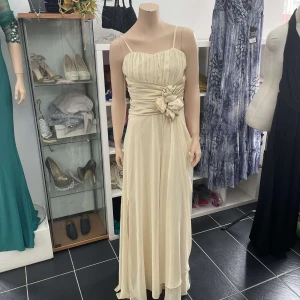 Champagnefärgad långklänning med rosettdetalj - Elegant långklänning i champagnefärg/beige med smala axelband och plisserad överdel. Markerad midja med bred drapering och stor rosett framtill. Klänningen har en mjuk, flytande kjol och vacker drapering även baktill. Perfekt för bal eller fest. Finns i storlek 36-42, erbjuder även att skräddarsy enligt dina egna preferenser! 🥰