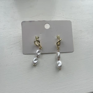 Guldiga örhängen med pärlor - Säljer ett par eleganta örhängen i guldton med två vita pärlor på varje örhänge. Örhängena har en snygg knutdetalj upptill och hängande pärlor som ger en stilren look.