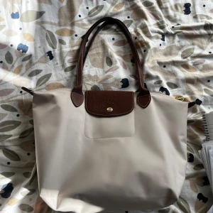 Beige axelväska från Longchamp Medium  - Säljer en klassisk beige axelväska från Longchamp med bruna handtag och lock i skinn. Väskan har en enkel design med guldknapp framtill och är perfekt för att bära med sig allt man behöver.