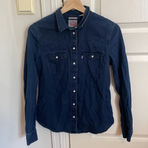 Mörkblå jeansskjorta från Levi's - Säljer en klassisk mörkblå jeansskjorta från Levi's med vita knappar och två bröstfickor med lock. Skjortan har lång ärm och tydliga sömmar som ger en snygg westernkänsla. Perfekt att styla med jeans eller chinos.