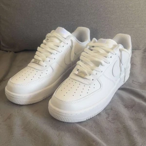 Nike Air Force 1 vita sneakers - Klassiska vita Nike Air Force 1 sneakers med låg profil och perforerad tå. Skorna har snörning, rund tå och platt sula. Perfekta för en clean och stilren look. Ikonisk Nike-logga på sidan och slitstark yttersula.
