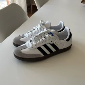 Adidas Samba sneakers i vitt och svart - Säljer ett par klassiska Adidas Samba sneakers i vitt med svarta ränder och grå mockadetaljer vid tån. Skorna är använda en gång och är i ett mycket fint skick! Ord pris 1 449kr.