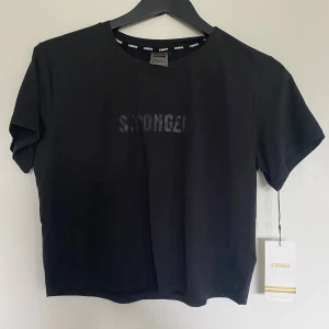 Svart croppad t-shirt från Stronger - Svart croppad t-shirt från Stronger med diskret logga framtill. T-shirten har korta ärmar och en loose passform, perfekt för en avslappnad stil. Tillverkad i mjuk bomull för extra komfort. Lappen kvar. Skicka gärna prisförlslag🩷