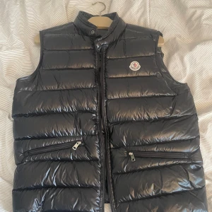 Svart dunväst från Moncler - Snygg svart dunväst från Moncler med glansig finish och ståkrage. Västen har två framfickor med dragkedja och Moncler-logga på bröstet. Perfekt för lager-på-lager och ger en sportig look. Modellen heter gui. Nästan helt oanvänd. Storlek 4 som motsvarar L. Nfc funkar se bild 4 för bevis 