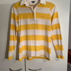 Gul- och vitrandig långärmad pikétröja från GANT - Säljer en klassisk långärmad pikétröja från GANT i gult och vitt med breda ränder. Tröjan har vit krage, knäppning vid halsen och broderad GANT-logga på bröstet. Perfekt för en sportig och avslappnad stil.