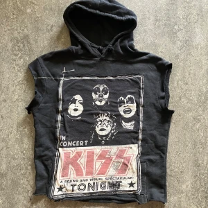 Svart KISS hoodie utan ärmar - Unik svart hoodie med tryck av KISS på både fram- och baksidan. Tröjan har huva och är ärmlös, perfekt för dig som gillar rock och vill sticka ut. Stort motiv på ryggen och konsertgrafik framtill. 