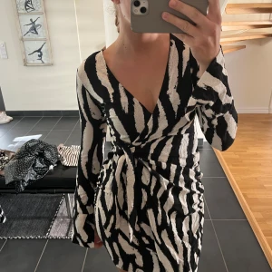Zebramönstrad omlottklänning med knytband - Snygg kort omlottklänning med zebramönster i svart och beige. Klänningen har lång ärm, djup v-ringning och markerad midja med knytband. 