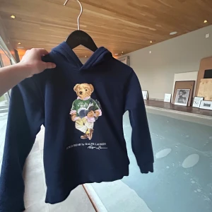 Mörkblå hoodie med björn från Polo Ralph Lauren - Säljer en mörkblå hoodie från Polo Ralph Lauren med den klassiska björnen på framsidan. Tröjan har huva och långa ärmar, perfekt för en cool och avslappnad stil. Ikonisk logga och mjukt material.