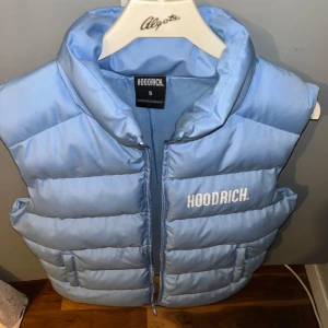 Ljusblå dunväst från Hoodrich - Säljer en ljusblå dunväst från Hoodrich med dragkedja och hög krage. Västen har två fickor framtill och tydlig Hoodrich-logga både fram och bak. Perfekt för lager-på-lager-stil under varma dagar. Lite använd inga skador finns o är väldigt ren