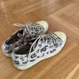 Beige sneakers med svart print från Acne Studios - Unika sneakers från Acne Studios med svart tecknat mönster över hela skon. Skorna har vita snören, rund tå och platt sula. Tåpartiet är vitt/ljusgult och märkesdetalj syns på plösen.