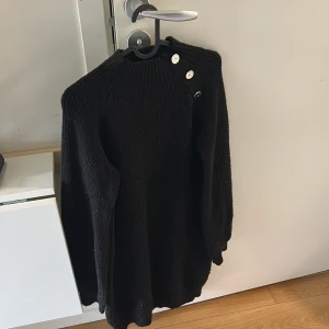 Svart stickad tröja med knappar - Mysig svart stickad klänning med lång ärm och dekorativa knappar vid axeln. Perfekt för kyligare dagar och enkel att matcha med olika outfits.