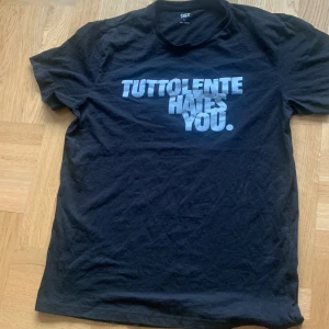 Svart t-shirt med tryck 'TUTTOLENTE HATES YOU.' - Cool svart t-shirt från Tuttolente med stort vitt tryck på bröstet där det står TUTTOLENTE HATES YOU. Vann den när dom kasta ut kläder. Storlek L men passar också M oversized. Hör av dig för fler frågor.