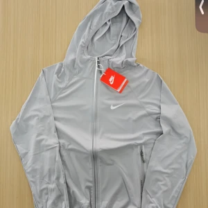 Grå Nike vindjacka - Nike vindjacka i ljusgrått med luva och dragkedja framtill. Jackan har lång ärm, två sidofickor med dragkedja och en diskret Nike-logga på bröstet. Perfekt för träning eller när du vill ha en sportig look. Den är storlek L men passar bättre som M