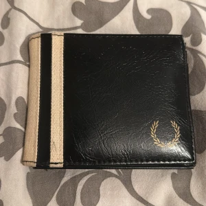 Svart och beige plånbok från Fred Perry - Säljer en stilren svart plånbok från Fred Perry med beige tygdetalj på sidan och klassisk lagerkrans-logga i guld. Plånboken har flera kortfack och ett fack med plastfönster för legitimation. Perfekt för dig som vill ha en snygg och praktisk accessoar.