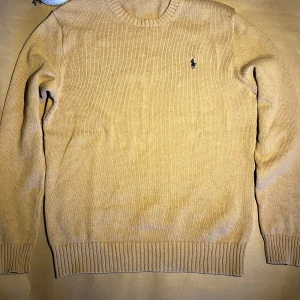 Beige stickad tröja från Polo Ralph Lauren - Säljer en klassisk beige stickad tröja från Polo Ralph Lauren med rund halsringning och diskret broderad logga på bröstet. Perfekt för lager-på-lager och enkel att matcha med det mesta.