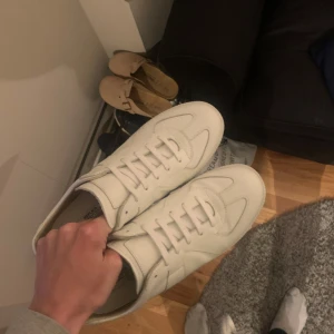 Vita sneakers från Maison Margiela - Säljer ett par stilrena vita sneakers från Maison Margiela. Skorna har klassisk design med rund tå, platt sula och snörning. Tillverkade i skinn med diskreta paneler på sidorna. Perfekta för dig som gillar minimalistisk och clean stil.