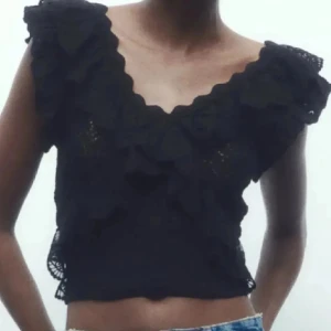 Svart virkad croppad topp med volang - Säljer en svart virkad croppad topp med volangdetaljer och v-ringning. Toppen har breda axelband och en romantisk känsla, perfekt till sommaren.