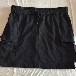 Svart kjol med fickor från Vero Moda - Svart kjol från Vero Moda med resår och dragsko i midjan. Kjolen har två stora fickor på sidorna och är i en mjuk bomullsblandning. Perfekt för en avslappnad och cool stil.