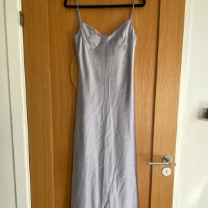 Ljusgrå, svagt lila långklänning från Zara - Säljer en elegant ljusgrå, svagt lila långklänning från Zara med smala axelband och lätt glansigt material. Klänningen har en v-ringad byst och öppen rygg med resårdetalj. Perfekt för sommarens speciella tillfällen, nypris 775kr.