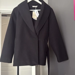 Svart kappa med bälte från Gina Tricot - Säljer en svart kappa från Gina Tricot i storlek XS. Jackan har ett brett slag, knyts med bälte i midjan och har långa ärmar. Aldrig använt.Säljet pågrund av att den inte kommer till användning. Hör av vid frågor eller fler bilder 🩷
