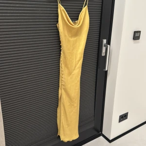 Gul långklänning i satin från Zara - Säljer en gul långklänning från Zara i glansigt satinmaterial. Klänningen har smala axelband, draperad urringning och dekorativa knappar längs sidan. Perfekt för dig som vill sticka ut med en elegant och trendig look.