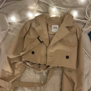 Beige croppad trenchcoat från Zara 140kr - Säljer en beige croppad trenchcoat från Zara med klassisk krage, dubbelknäppning och bälte i midjan. Använd fåtal gånger. Strl M. 140kr